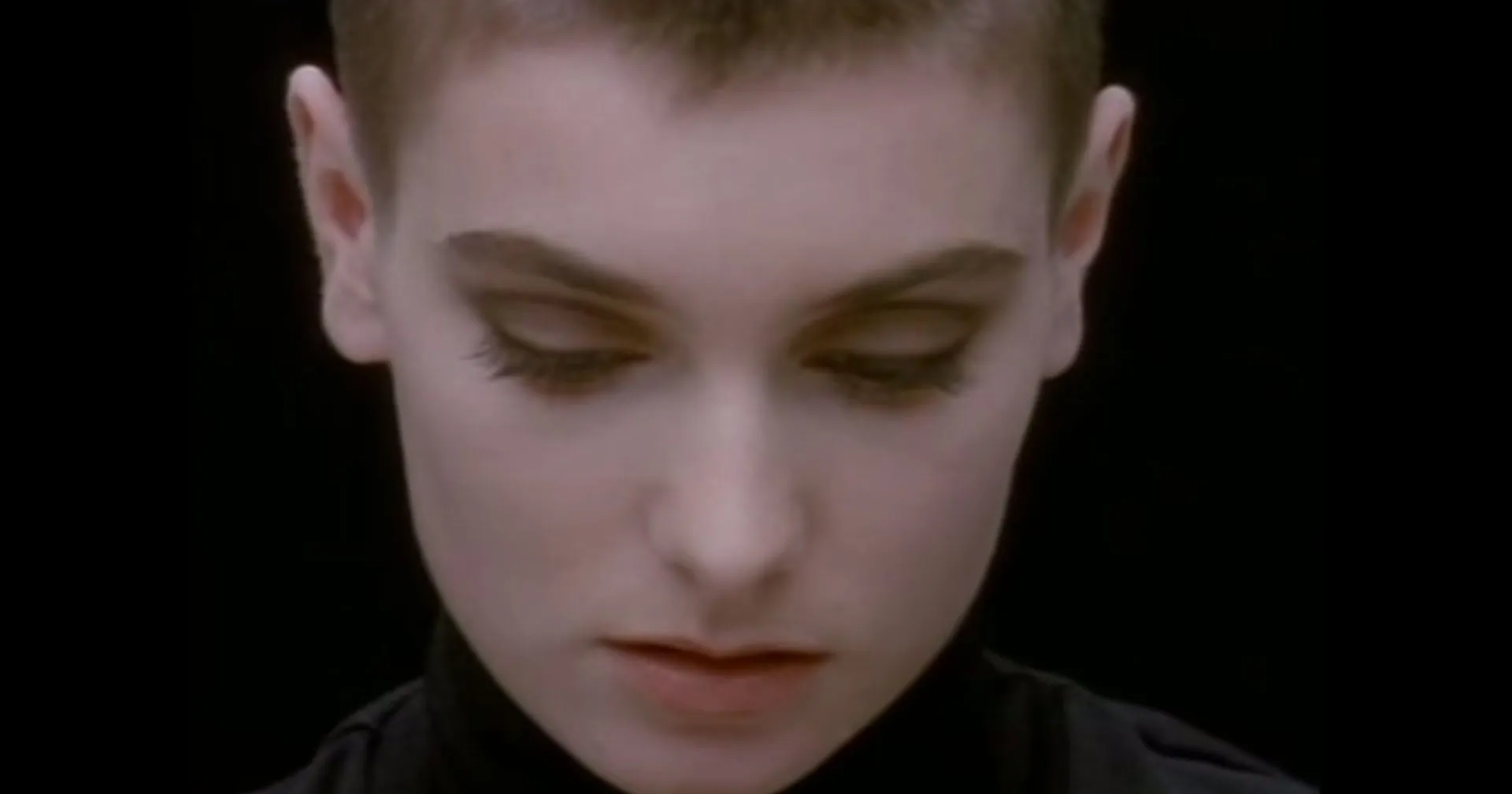 Zbliżenie twarzy Sinéad O'Connor z zamkniętymi oczami podczas kultowego klipu do piosenki 'Nothing Compares 2 U'. Ciemne tło podkreśla emocjonalny i melancholijny wyraz twarzy artystki, symbolizujący temat tęsknoty i straty.